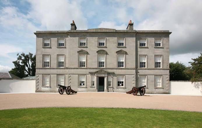 The grey stone Georgian facade of Oldbridge House, with two historic cannons on the gravel forecourt and a lawn in front.
